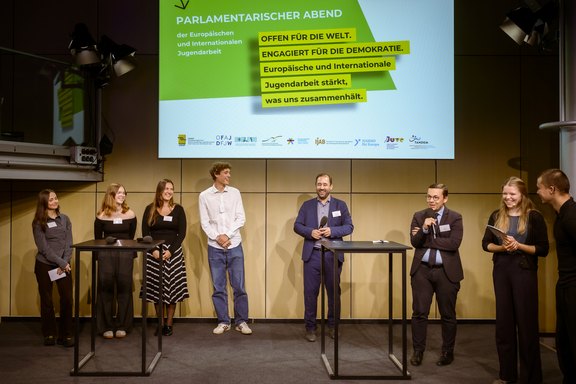 Vier junge Menschen und zwei Abgeordnete stehen zusammen mit zwei Moderator:innen bei einer Fragerunde auf einer Bühne.